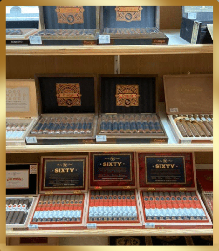 A display of cigar boxes on wooden shelves. The top shelf shows two open black boxes with gold accents and cigars. The middle shelf features more open boxes with cigars, and the bottom shelf has three open 'Rocky Patel Sixty' cigar boxes, with cigars arranged in rows of red and white bands.