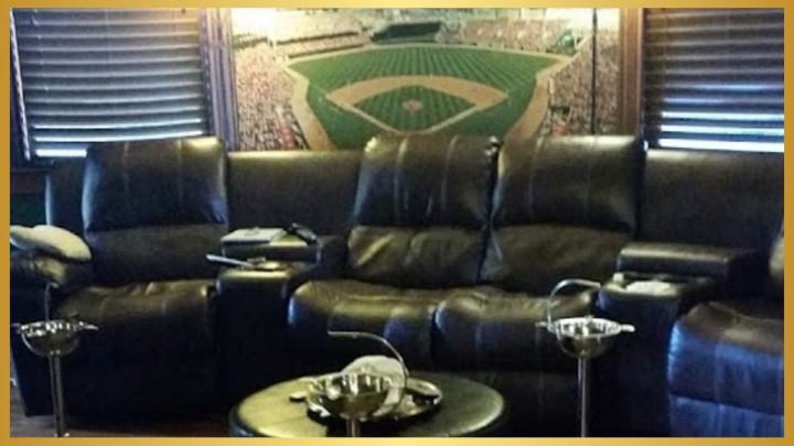 A cozy lounge area with dark leather recliners facing a large artwork of a baseball field behind a wooden blind, featuring ash trays and side tables.