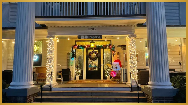 The brightly lit entrance of cigar mansion, adorned with white string lights, wreaths, and festive holiday decorations, including a snowman figure, on a porch with large white pillars.