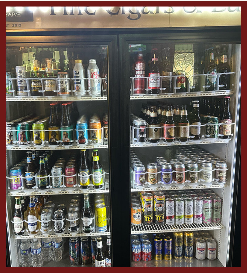 A large glass-door refrigerator filled with numerous cans and bottles of beer and other beverages, organized on multiple shelves.