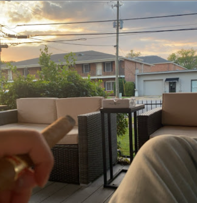 A close-up, first-person view of a hand holding a lit cigar, with a blurred outdoor patio setting in the background including a cushioned outdoor sofa and a table, under a sky with visible clouds and a sunset glow.