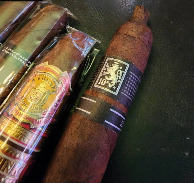 A close-up shot of two cigars. The cigar in the foreground has a dark brown wrapper and a black and white band with '10' and a crest. Behind it, another cigar in clear plastic packaging has a gold and red band with 'FUENTE' visible.