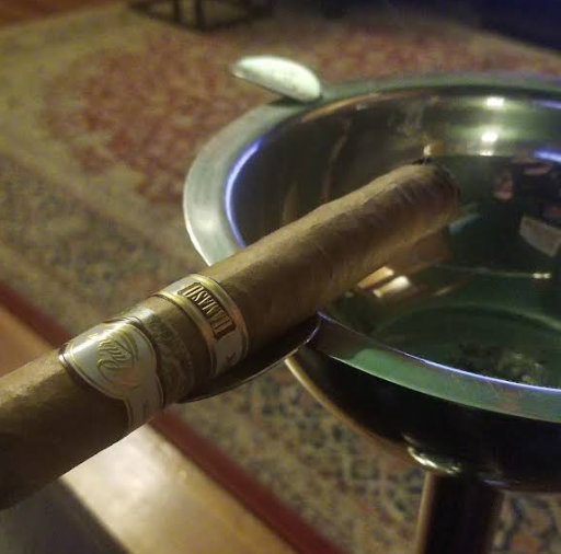 A close-up shot of a light-colored cigar resting on the edge of a shiny silver ashtray. The cigar has a white and gold band. A blurred patterned rug is visible in the background.