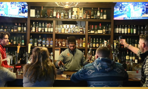 A lively bar scene with a well-stocked liquor shelf in the background. Several people are seated at the bar, interacting with a bartender. A TV screen is visible above the bar.