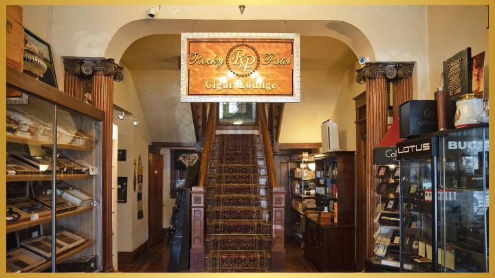 The interior of a cigar lounge with a grand staircase leading to an upper floor. Display cases filled with cigars line the walls, and a 'Rocky's Place Cigar Lounge' banner hangs over the staircase.