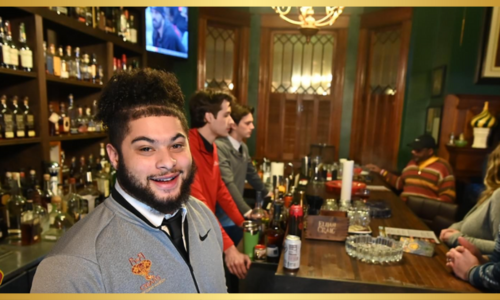 A young man with curly hair smiling and looking towards the left, standing in front of a bar area with liquor bottles on shelves and other patrons in the background. A TV screen is also visible.